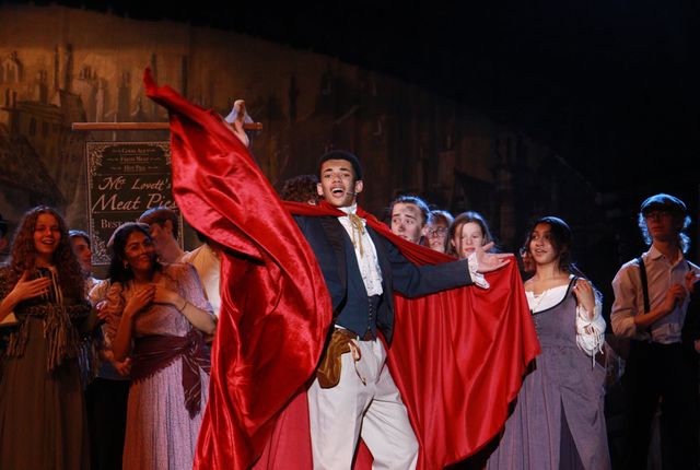 Pirelli sweeps on in a red cape in front of the ensemble and Mrs Lovett's pie shop sign